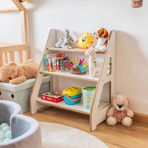 Pode incluir: Uma estante de madeira branca com tr&ecirc;s prateleiras, cheia de brinquedos infantis. Os brinquedos incluem bichos de pel&uacute;cia, livros e brinquedos coloridos. A estante est&aacute; em um quarto infantil com um tapete e outros brinquedos.