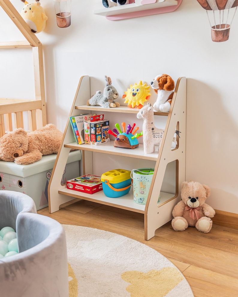 Pode incluir: Uma estante de madeira branca com tr&ecirc;s prateleiras, cheia de brinquedos infantis. Os brinquedos incluem bichos de pel&uacute;cia, livros e brinquedos coloridos. A estante est&aacute; em um quarto infantil com um tapete e outros brinquedos.