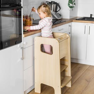 May include: A light-coloured wooden learning tower with a tall, rectangular frame, side cutouts for handles, and a built-in step. The structure features a smooth, natural wood grain finish, a sturdy base, and a horizontal bar across the top back section.
