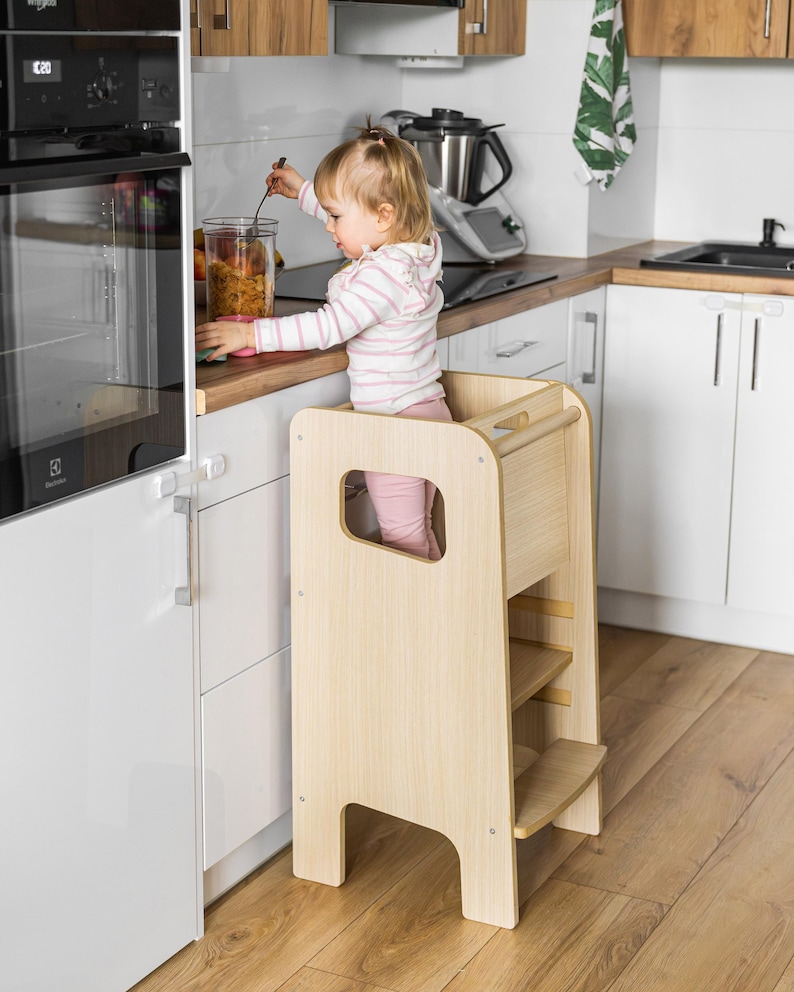 May include: A light-coloured wooden learning tower with a tall, rectangular frame, side cutouts for handles, and a built-in step. The structure features a smooth, natural wood grain finish, a sturdy base, and a horizontal bar across the top back section.