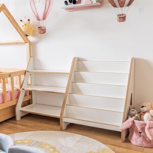 Pode incluir: Duas estantes brancas e em tons de madeira com v&aacute;rias prateleiras num quarto infantil. O quarto apresenta um tapete redondo com um design de arco-&iacute;ris, uma cama pequena e decalques de parede de bal&otilde;es de ar quente. Animais de pel&uacute;cia colocados nas prateleiras e numa cesta rosa.