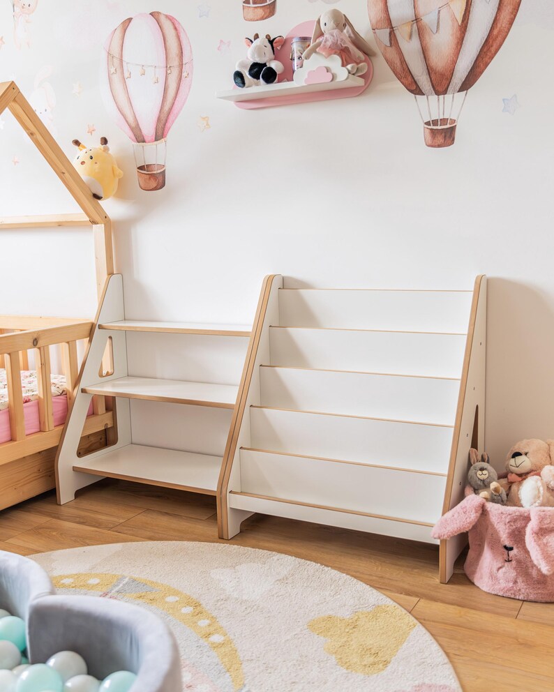 Pode incluir: Duas estantes brancas e em tons de madeira com v&aacute;rias prateleiras num quarto infantil. O quarto apresenta um tapete redondo com um design de arco-&iacute;ris, uma cama pequena e decalques de parede de bal&otilde;es de ar quente. Animais de pel&uacute;cia colocados nas prateleiras e numa cesta rosa.