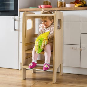 May include: A light-coloured wooden learning tower with a safety rail, a standing platform, and a bottom step. The structure features smooth, natural wood grain surfaces, vertical side panels with cut-out handles, and a sturdy frame design.