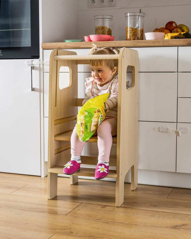 May include: A light-coloured wooden learning tower with a safety rail, a standing platform, and a bottom step. The structure features smooth, natural wood grain surfaces, vertical side panels with cut-out handles, and a sturdy frame design.