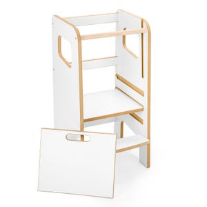 May include: A white and light wood learning tower with a tall frame, a platform, and a horizontal bar at the top. A separate white rectangular board with a cutout handle is positioned in front of the tower, which features side cutouts.