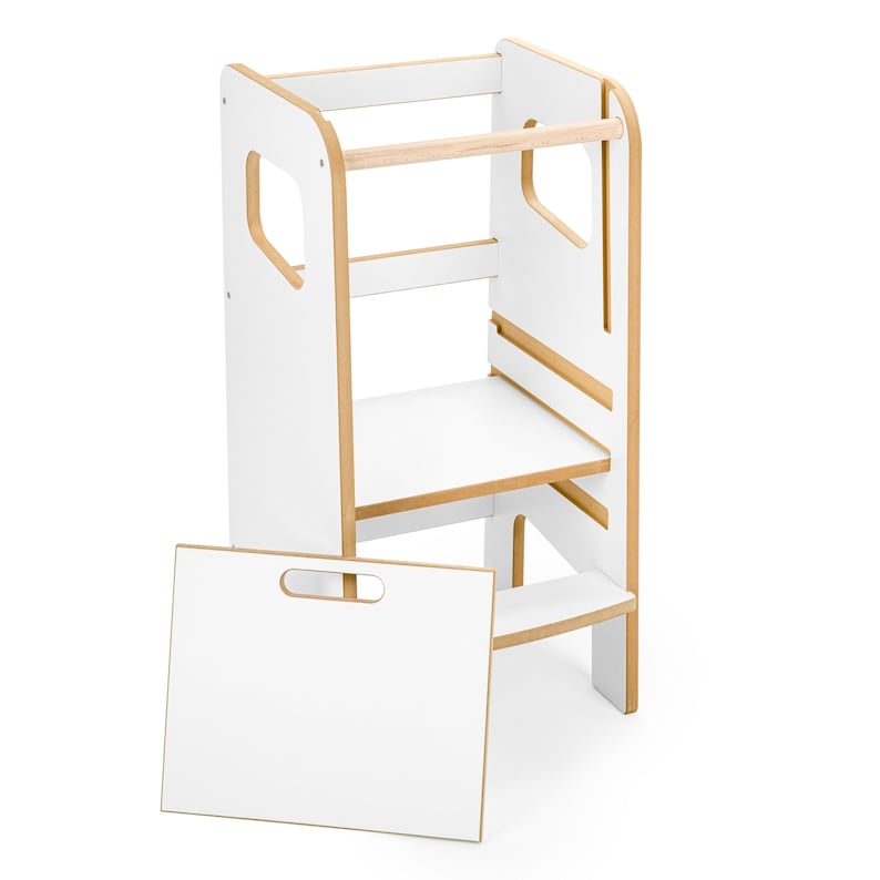 May include: A white and light wood learning tower with a tall frame, a platform, and a horizontal bar at the top. A separate white rectangular board with a cutout handle is positioned in front of the tower, which features side cutouts.