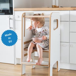 May include: A white and light wood kitchen learning tower with a safety rail and a step, featuring a blue circular badge with the text Made in EU. The structure has side cutouts and a stable base, designed to provide elevation for a small child.