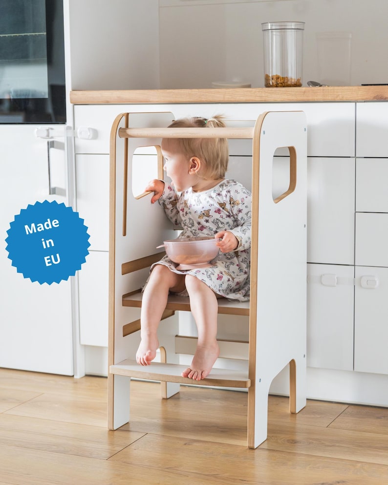 May include: A white and light wood kitchen learning tower with a safety rail and a step, featuring a blue circular badge with the text Made in EU. The structure has side cutouts and a stable base, designed to provide elevation for a small child.