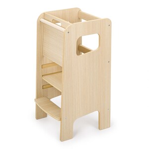 May include: A light-coloured wooden learning tower featuring a tall, open-frame design with a sturdy base, a middle platform, and a top safety rail. The structure includes side cutouts for handles and is constructed from smooth, light-toned wood panels.