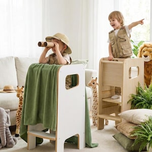 May include: Two wooden learning towers, one white and one natural wood finish, featuring side cutouts and steps. The white tower is draped with a solid green blanket, while the natural wood tower stands beside it, both designed for children to reach elevated surfaces.