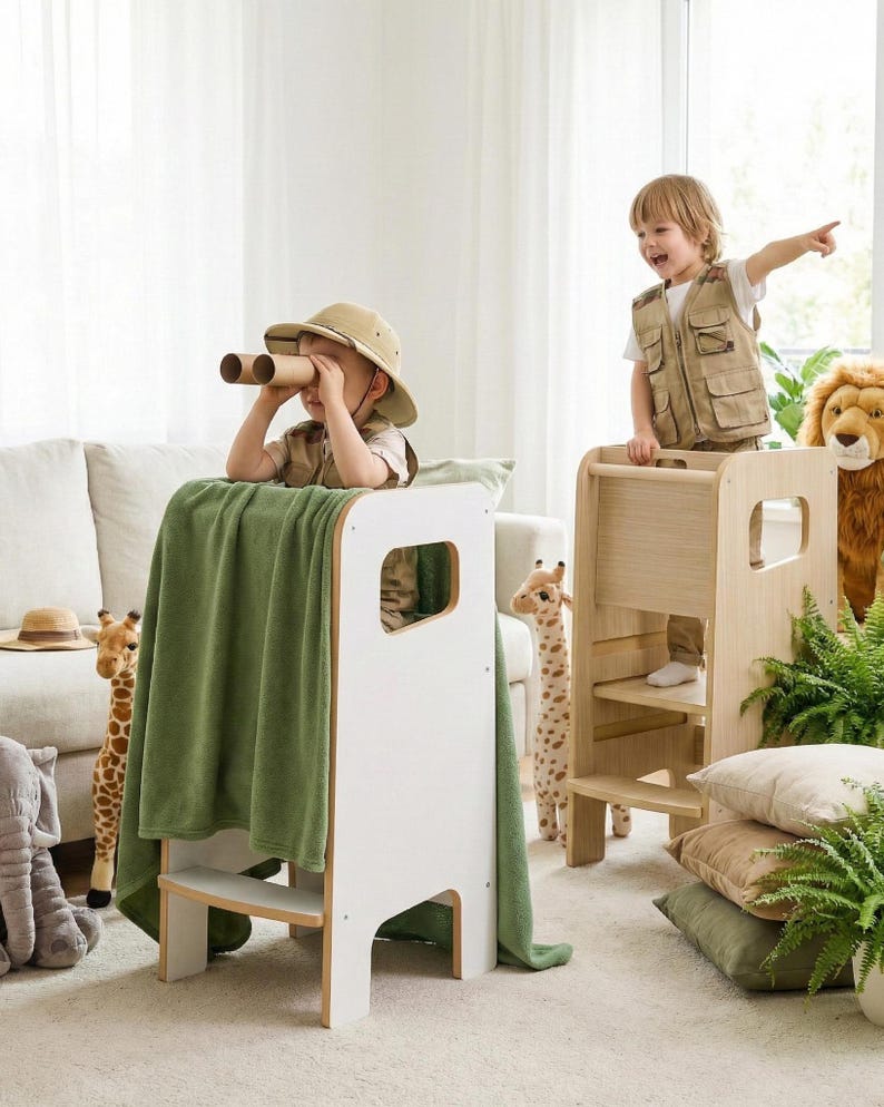 May include: Two wooden learning towers, one white and one natural wood finish, featuring side cutouts and steps. The white tower is draped with a solid green blanket, while the natural wood tower stands beside it, both designed for children to reach elevated surfaces.