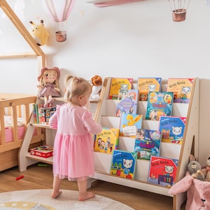 Pode incluir: Um quarto infantil com uma estante branca cheia de livros infantis coloridos. A estante fica ao lado de uma estrutura de cama de madeira. Bal&otilde;es de ar quente decorativos e bichos de pel&uacute;cia adornam as paredes. Uma crian&ccedil;a pequena com um vestido rosa est&aacute; em frente &agrave; estante.