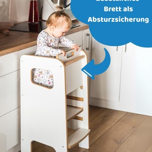 May include: A white wooden learning tower with a natural wood trim, featuring a safety board at the top. The structure includes a step and a platform for standing. A blue speech bubble graphic with the text "Zus&auml;tzliches Brett als Absturzsicherung" points to the safety board.