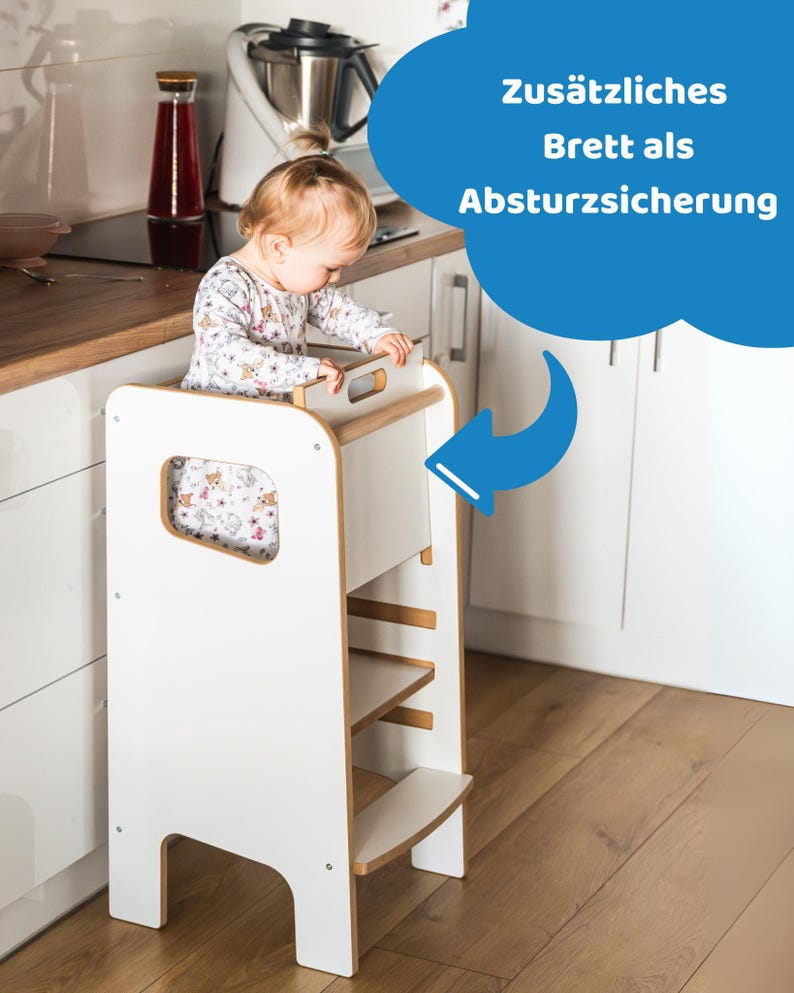 May include: A white wooden learning tower with a natural wood trim, featuring a safety board at the top. The structure includes a step and a platform for standing. A blue speech bubble graphic with the text "Zus&auml;tzliches Brett als Absturzsicherung" points to the safety board.