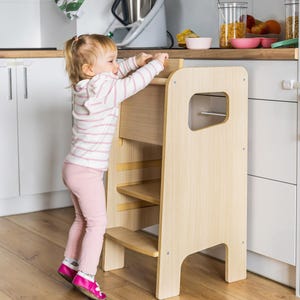 May include: A light-coloured wooden learning tower with a step stool design, featuring side panels with cutout handles and two horizontal steps. The structure is made of smooth, light-toned wood and provides a stable platform for reaching worktops.