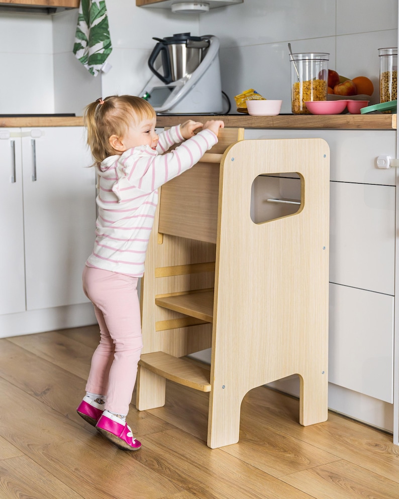 May include: A light-coloured wooden learning tower with a step stool design, featuring side panels with cutout handles and two horizontal steps. The structure is made of smooth, light-toned wood and provides a stable platform for reaching worktops.