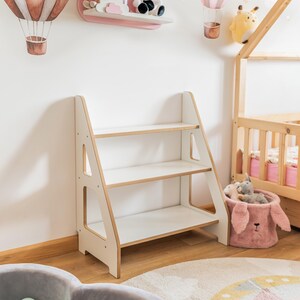 Pode incluir: Uma estante de madeira branca e castanho claro com tr&ecirc;s prateleiras. A estante tem um design semelhante a uma escada e est&aacute; colocada no quarto de uma crian&ccedil;a. O quarto tamb&eacute;m apresenta uma prateleira rosa com bichos de pel&uacute;cia, uma estrutura de cama em forma de casa de madeira e uma piscina de bolinhas cinza.