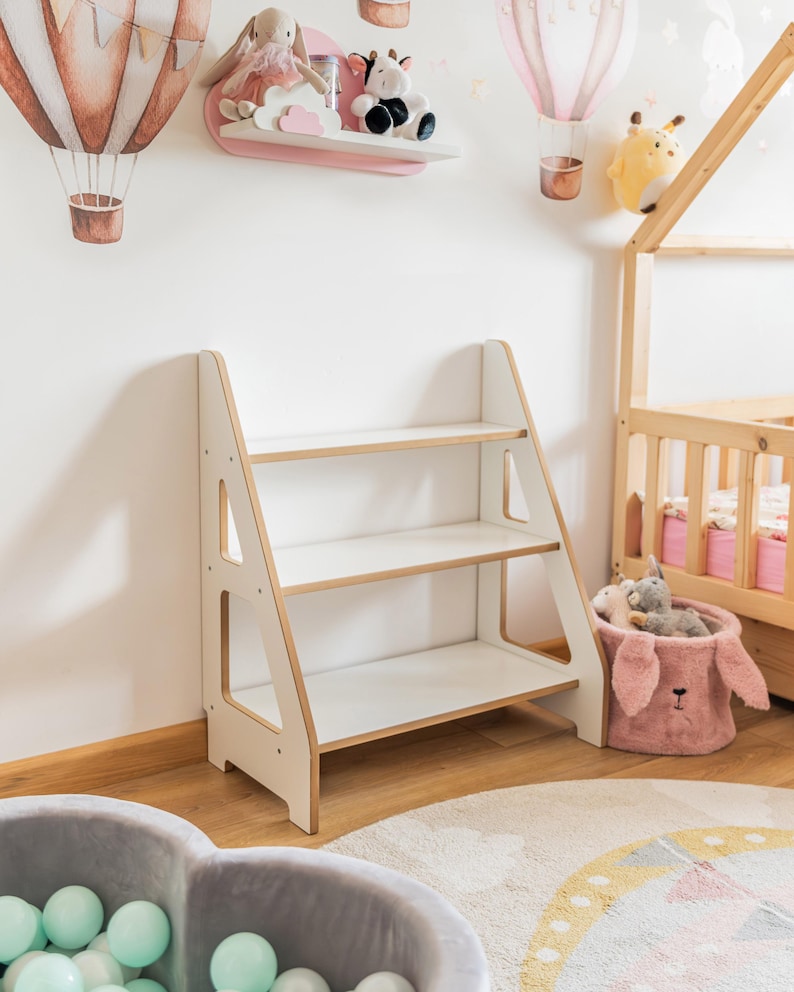 Pode incluir: Uma estante de madeira branca e castanho claro com tr&ecirc;s prateleiras. A estante tem um design semelhante a uma escada e est&aacute; colocada no quarto de uma crian&ccedil;a. O quarto tamb&eacute;m apresenta uma prateleira rosa com bichos de pel&uacute;cia, uma estrutura de cama em forma de casa de madeira e uma piscina de bolinhas cinza.