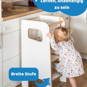 May include: A white and natural wood learning tower with a wide step, featuring side cutouts and a top safety rail. The tower includes blue text overlays that read "Sehr leichter Zugang", "Lernen, unabh&auml;ngig zu sein", and "Breite Stufe".