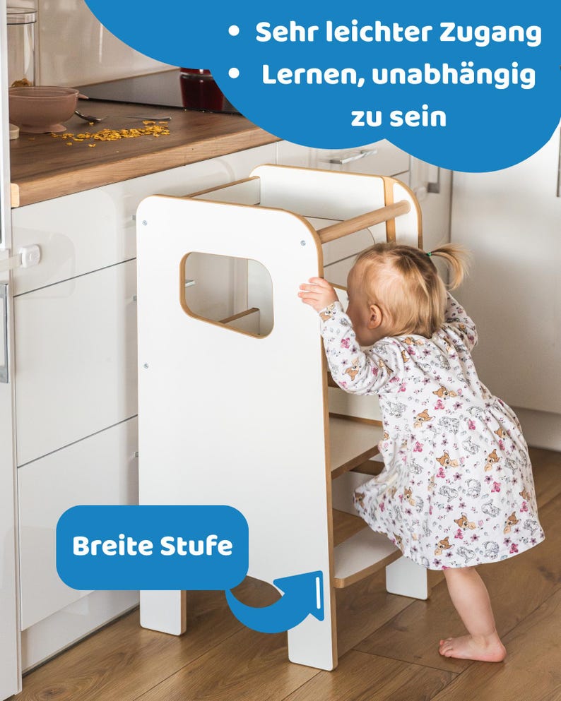 May include: A white and natural wood learning tower with a wide step, featuring side cutouts and a top safety rail. The tower includes blue text overlays that read "Sehr leichter Zugang", "Lernen, unabh&auml;ngig zu sein", and "Breite Stufe".