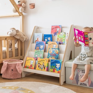 Pode incluir: Uma estante de madeira branca cheia de livros infantis coloridos. Uma crian&ccedil;a pequena est&aacute; lendo um livro com capa rosa. O quarto tem um tema l&uacute;dico com bichos de pel&uacute;cia e um tapete decorativo.