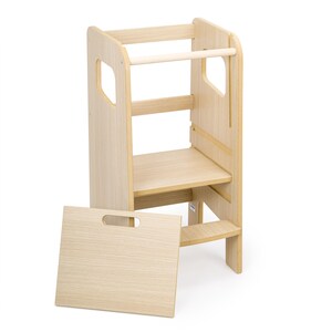 May include: A light-coloured wooden learning tower with a platform, a safety bar, and side cutouts for handles. A separate rectangular wooden panel with a handle cutout is positioned in front of the tower, featuring a light wood grain finish.