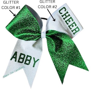 May include: Green and white cheer bow with glitter. The bow has the word "CHEER" on one side and the name "ABBY" on the other side.
