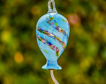 May include: A blue glass hummingbird feeder with a red feeding tube. The feeder is decorated with colorful swirls and speckles.