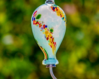 May include: A blue glass hummingbird feeder with a colorful, speckled design. The feeder has a long, thin spout and a red tip.