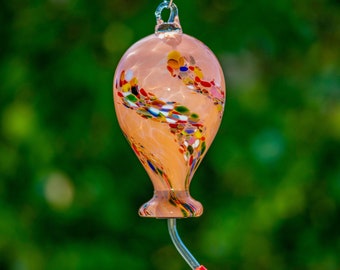 Könnte beinhalten: Ein Glas-Kolibri-Futterspender mit einem pfirsichfarbenen Sockel und einem wirbelnden Muster aus bunten Glasperlen. Der Futterspender hat einen roten Futteranschluss und ein klares Plastikrohr.