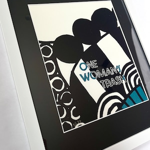 Op de afbeelding: Zwart-witte abstracte kunstprint met de tekst "ONE WOMAN'S TRASH" in tealblauw. De print is ingelijst in een wit frame.