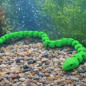 Op de afbeelding: Een felgroen, beweeglijk slangen speelgoed ligt op een bedje van kleine, veelkleurige kiezelstenen. De speelgoedslang bevindt zich in een gebogen positie, met zijn kop en staart zichtbaar. De achtergrond toont waterplanten en een blauwgroen watereffect.