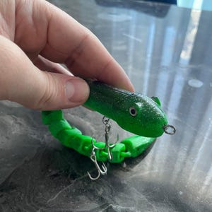 Puede incluir: Un señuelo de pesca verde y negro con forma de serpiente. El señuelo tiene un cuerpo segmentado y dos anzuelos plateados. El señuelo está sujeto por una mano.