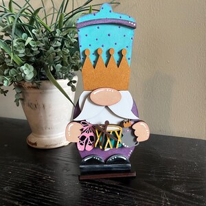 May include: A wooden gnome figurine with a purple hat, a blue and white crown, and a white beard. The gnome is holding a drum, a drumstick, and a small mouse figurine.