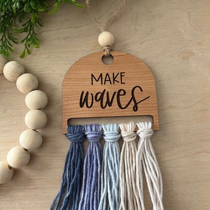 May include: Wooden sign with the text "Make Waves" in a cursive font. The sign has a light wood finish and is decorated with a fringe of blue, light blue, and white yarn.