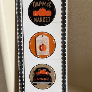 May include: A white wooden sign with a black frame and three round decorations. The top decoration says "Harvest Market" with three orange pumpkins. The middle decoration says "Pumpkin Patch" with a single orange pumpkin. The bottom decoration says "hello fall" with a black truck carrying pumpkins.