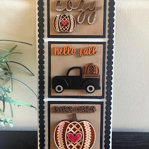 May include: A wooden sign with three panels featuring fall-themed designs. The top panel says "Stay Cozy" with a pumpkin and heart. The middle panel says "Hello Fall" with a truck carrying a pumpkin. The bottom panel says "Sweater Weather" with a pumpkin and heart.