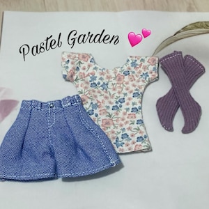 Pode incluir: Um traje de boneca em miniatura com uma saia jeans, um top floral e meias roxas. O top tem um fundo branco com estampas florais rosa e azul. O texto "Pastel Garden" está escrito em itálico.