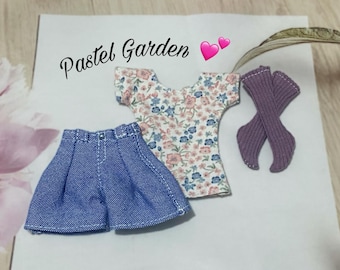 Peut inclure: Une tenue de poupée miniature comprenant une jupe en jean, un haut floral et des chaussettes violettes. Le haut a un fond blanc avec des motifs floraux roses et bleus. Le texte "Pastel Garden" est écrit en cursive.