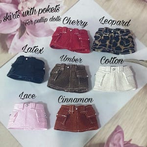 Peut inclure: Mini-jupes pour poupées de différentes couleurs et motifs, dont rouge, léopard, noir, marron, blanc, rose et rouille. Chaque jupe a des poches et est étiquetée avec des noms de tissus comme "Cherry" et "Cotton".