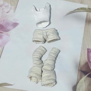 May include: A miniature doll clothing set, featuring a white ribbed tank top, light gray shorts, and matching leg warmers. The set is made of a soft, textured fabric. The items are arranged on a white surface, with floral accents in the background.