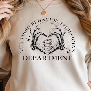 Op de afbeelding: Een beige sweatshirt met de tekst "The Tired Behavior Technician Department" gedrukt op de voorkant. De tekst wordt omringd door een zwart-witte illustratie van een skelet dat een stapel koffiekopjes vasthoudt.
