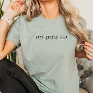 May include: Sage green t-shirt with the phrase "it's giving BCBA" printed in black. The shirt has short sleeves and a crew neck. The text is centered on the chest.