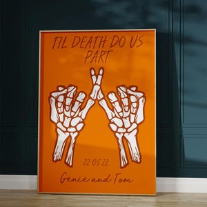 Op de afbeelding: Ingelijste oranje print met witte skelet handen die een vredesteken vormen. De tekst "TIL DEATH DO US PART" staat bovenaan, met de datum "22 05 22" en de namen "Genie and Tom" onderaan.