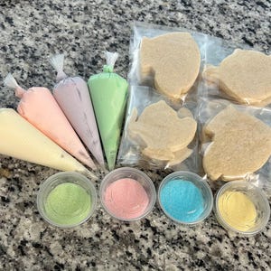 May include: A set of cookie decorating supplies, including four bags of icing in yellow, pink, purple, and green, four small containers of sprinkles in green, pink, blue, and yellow, and six sugar cookies in the shape of fish.