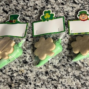 May include: Three green and white bags with a shamrock cookie inside. Each bag has a different topper with a leprechaun, a shamrock, and a hat.