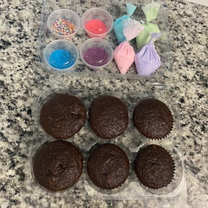 May include: A clear plastic container with six chocolate cupcakes and a separate container with sprinkles, sugar, and frosting in various colors.