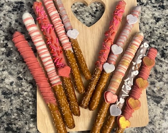 May include: A wooden board with a heart-shaped cutout holds a collection of chocolate-covered pretzels decorated with pink, white, and gold sprinkles and heart-shaped candies.