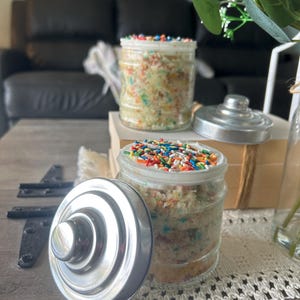 May include: Two glass jars filled with cake and topped with colorful sprinkles. The jars have silver lids and sit on a table with a decorative doily. One lid is off, revealing the cake inside. The cake has colorful sprinkles.