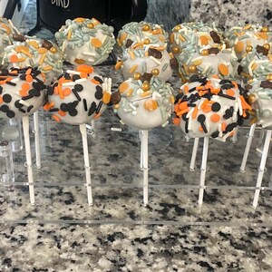 May include: A display of white cake pops decorated with various sprinkles, including black, orange, and gold. The cake pops are arranged on a clear acrylic stand.